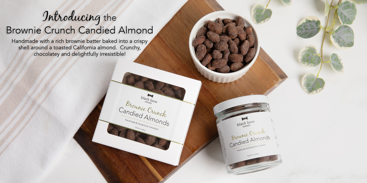 A picture of Black Bow Sweets Brownie Crunch Candied Almonds in a jar, a box and a small bowl on top of a wooden cutting board with a towel across it. 
 There are a few green leaves in the right corner and text in the left corner saying "Introducing the Brownie Crunch Candied Almond. Handmade with a rich brownie batter baked into a crispy shell around a toasted California almond. Crunchy, chocolatey and delightfully irresistible!